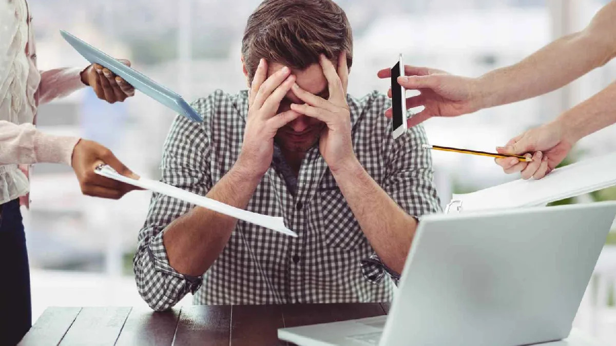 Homem sentado em frente ao computador, cercado por colegas entregando papéis e celulares, demonstrando sobrecarga de trabalho prejudicial à saúde mental.