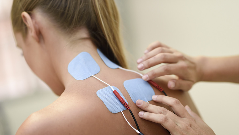 Aplicação de eletroestimulação muscular em paciente com lesão na lombar, com eletrodos retangulares fixados na região cervical e lombar, conectados a fios de condução elétrica para tratamento fisioterapêutico.