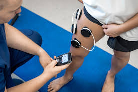 Aplicação de estimulação elétrica em paciente com lesão na coxa, utilizando eletrodos circulares fixados na parte frontal da coxa e equipamento de controle manual operado pelo fisioterapeuta.