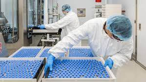 Linha de produção mostrando como é feita a produção de medicamentos, com equipamentos automatizados e operadores com vestimenta esterilizada.