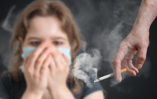 Criança usando máscara facial e cobrindo o nariz com as mãos, enquanto um adulto próximo segura um cigarro aceso, com fumaça visível se espalhando no ar.